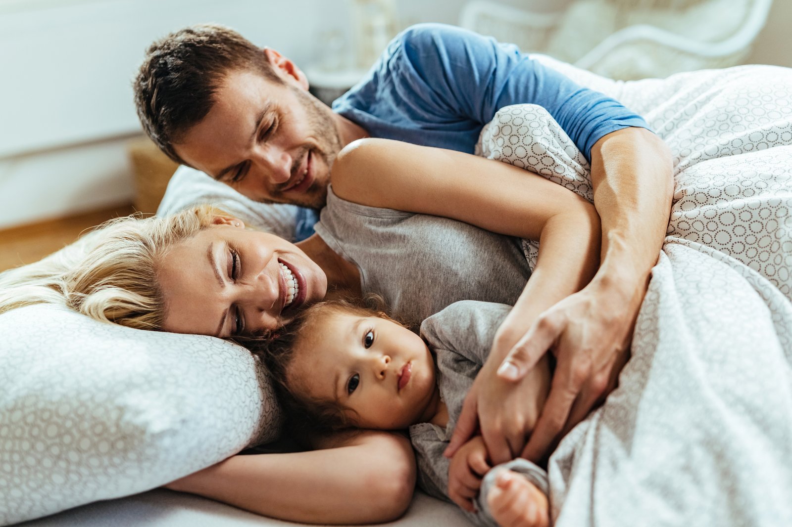 Junge Familie liegt eng umschlungen im Bett — Vater, Mutter und Kleinkind kuscheln am Morgen