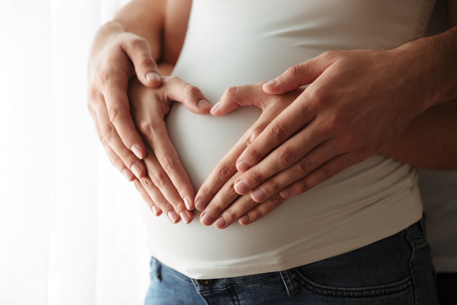 Hände eines Paares formen ein Herz auf dem Bauch der Frau — Hoffnung und Verbindung in der frühen Schwangerschaft